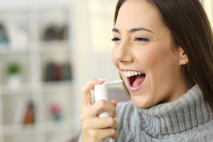 Happy woman wearing jersey using a analgesic spray to soften the throat sitting on a sofa in a house interior